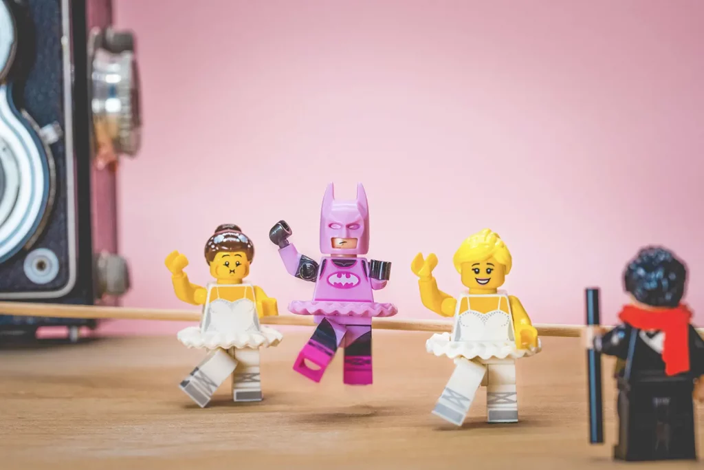 Cette image sur le thème de la Legography représente trois Lego qui prenne un cours de danse Classic. L'un dés Lego est Batman déguisé en Rose.